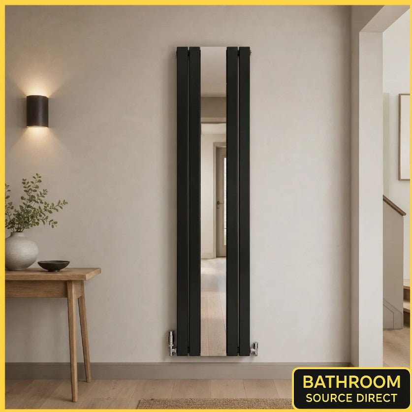A tall, narrow black radiator with an integrated mirror, mounted on a beige wall in a modern bathroom setting. A wooden side table with a vase and plant is visible on the left, and a hallway is visible through the mirror and on the right.